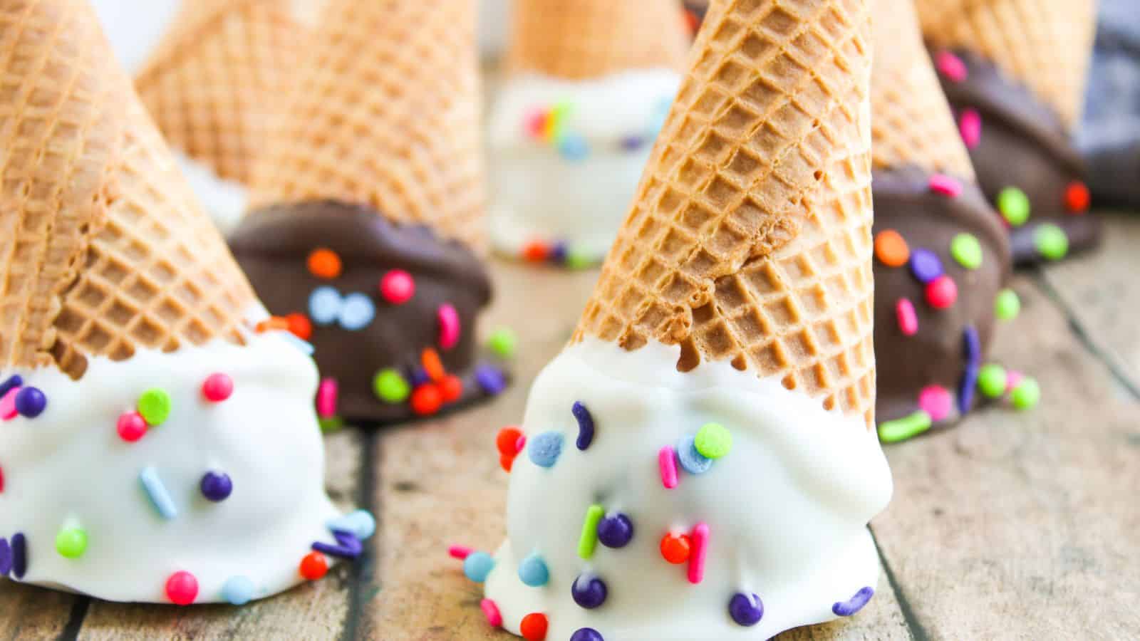Waffle ice cream cones dipped in white and dark chocolate, decorated with colorful sprinkles, arranged on a wooden surface.