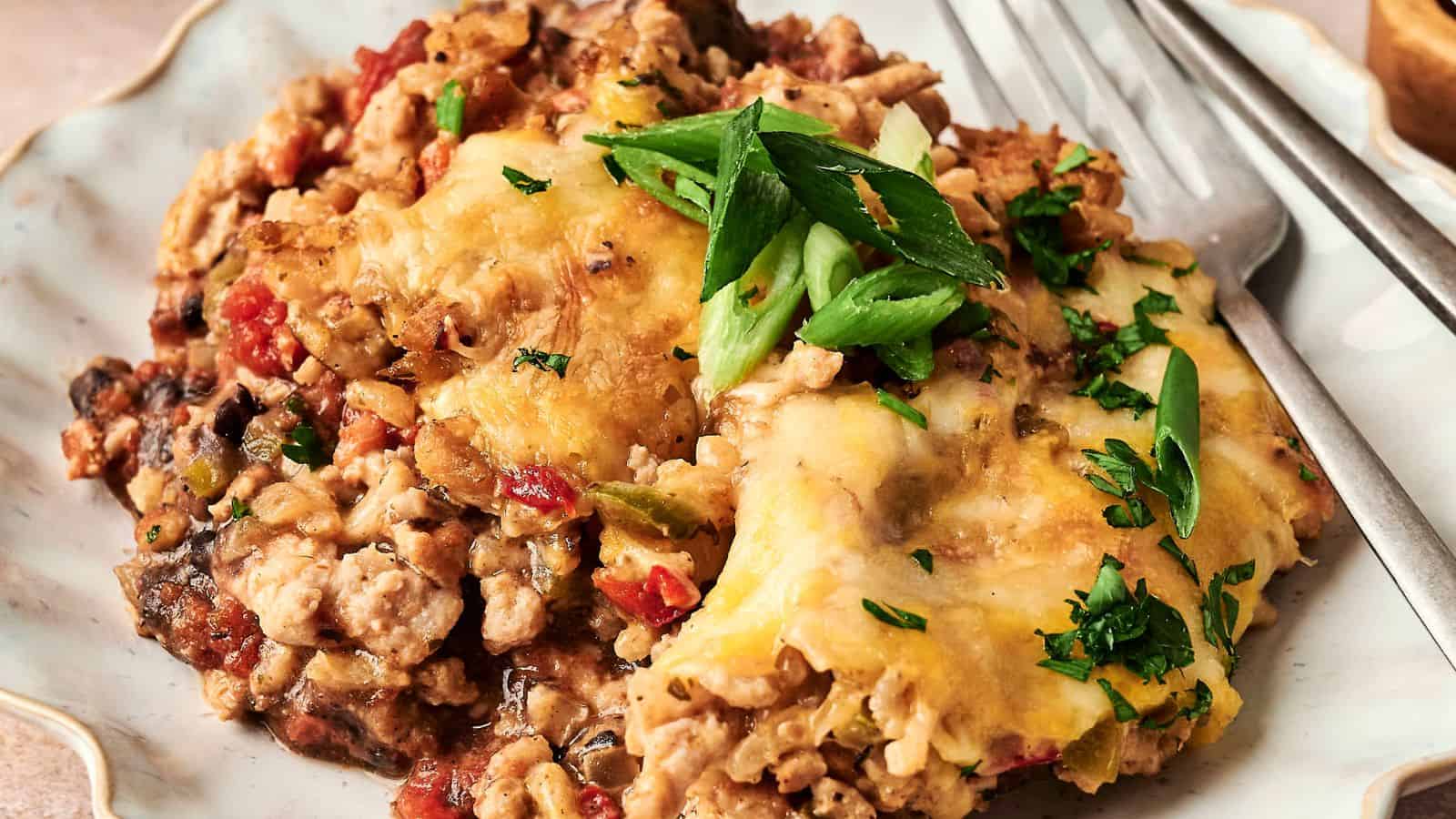A plated serving of cheesy casserole with ground meat, topped with sliced green onions and parsley, accompanied by a fork.