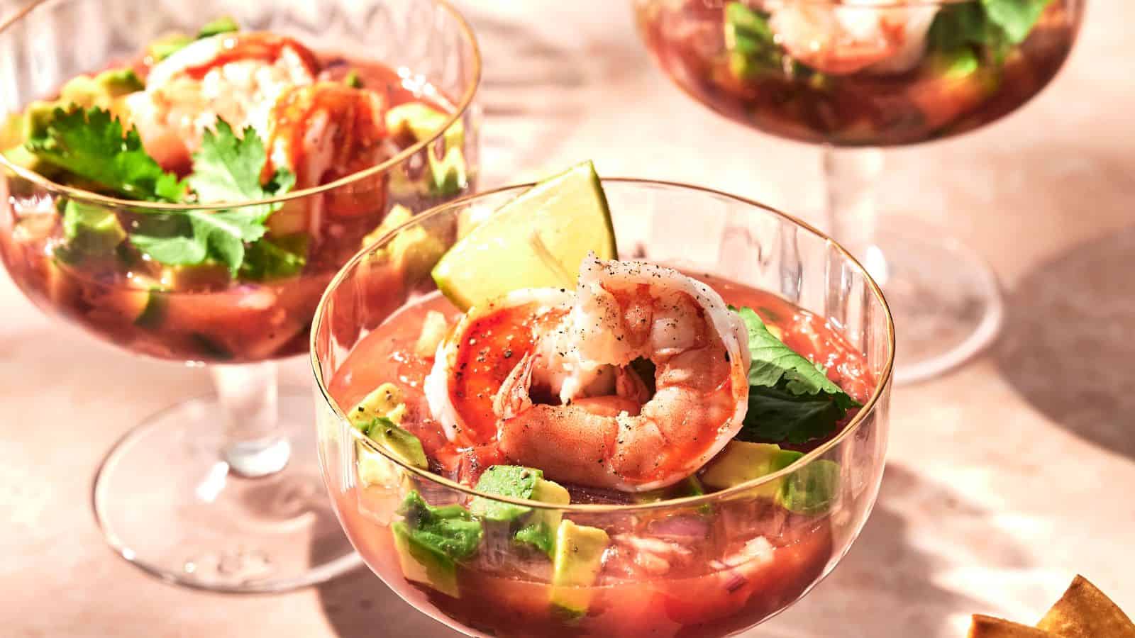 Three glasses of shrimp cocktail with avocado, lime wedge, and cilantro garnish in a tomato-based sauce, placed on a light-colored surface.