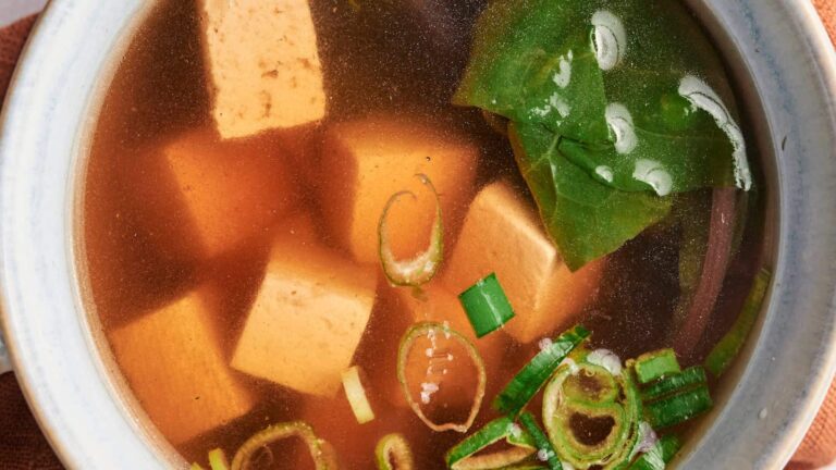 A bowl of miso soup with tofu cubes, sliced green onions, and leafy greens in a clear broth.
