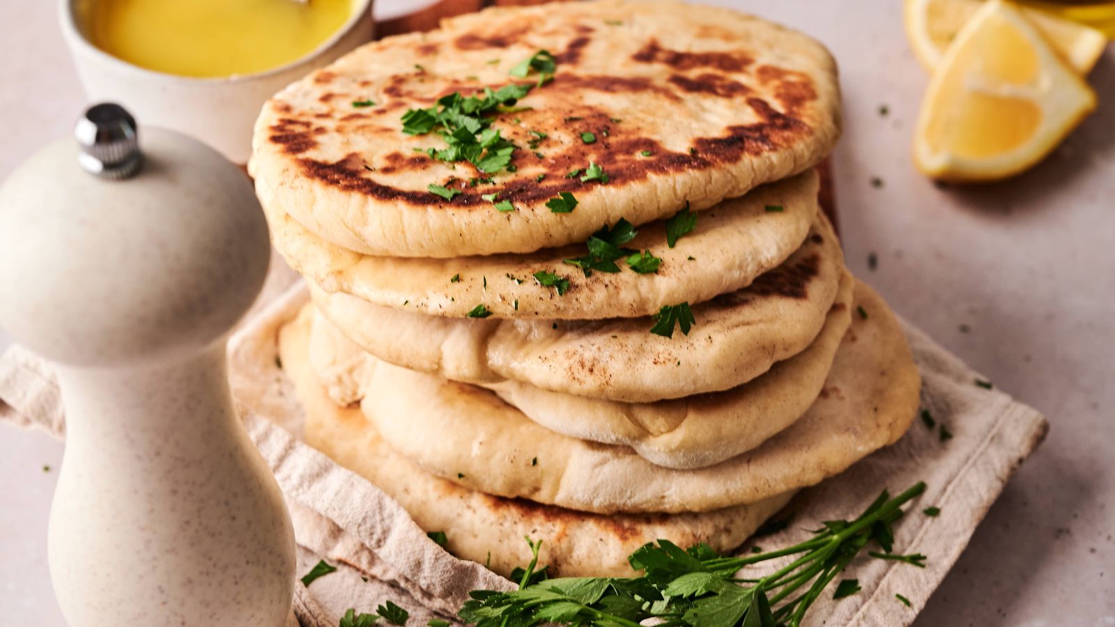 A stack of flatbreads garnished with chopped parsley sits on a cloth, with a white pepper grinder, fresh herbs, a lemon wedge, and a bowl of yellow sauce nearby.