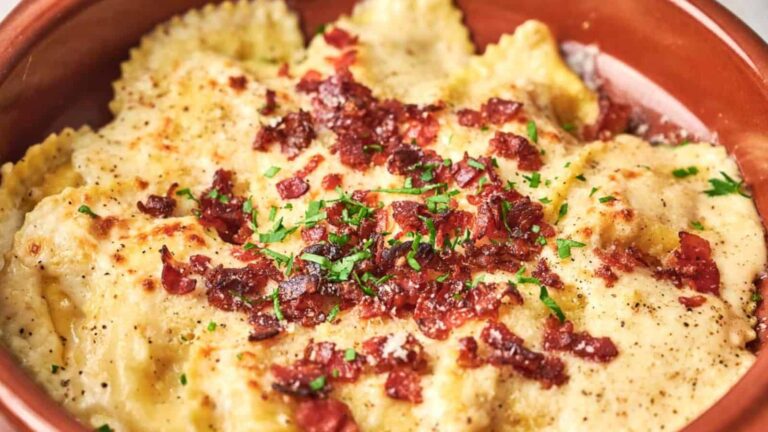 A bowl of ravioli topped with creamy sauce, crispy bacon bits, chopped parsley, and black pepper.