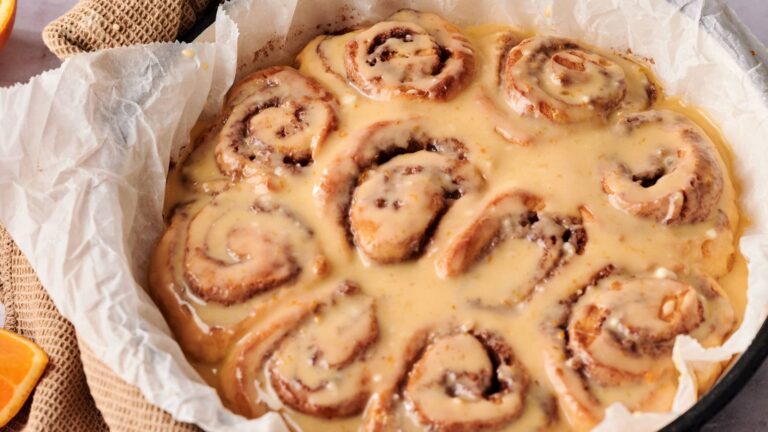 A round baking dish lined with parchment paper holds freshly baked cinnamon rolls covered in a thick layer of icing.
