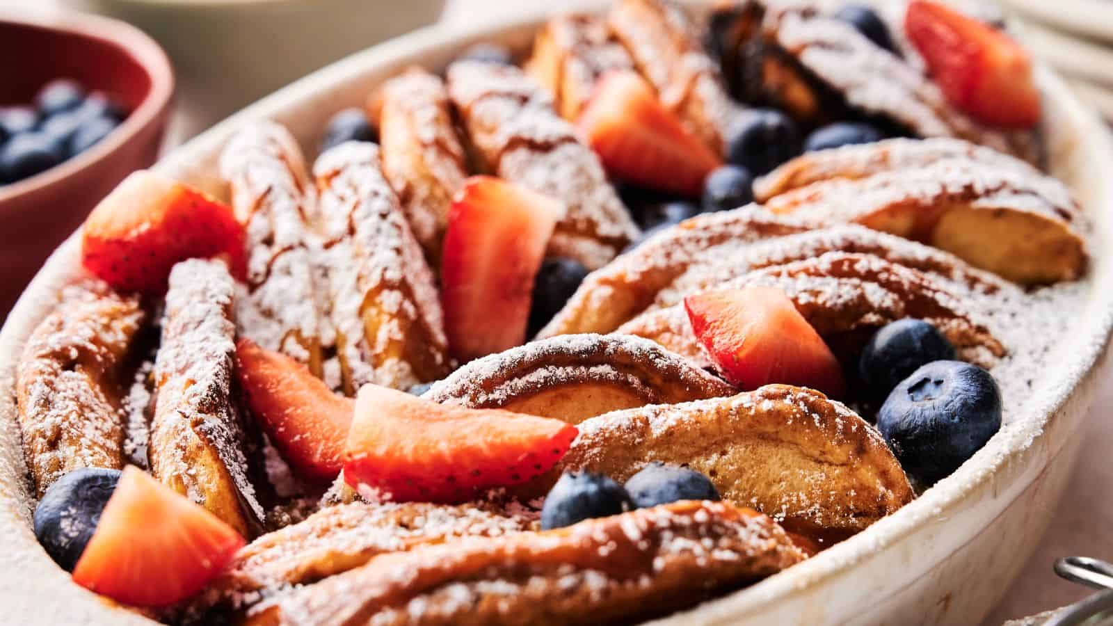 A baked French toast casserole topped with powdered sugar, sliced strawberries, and blueberries in a white dish.