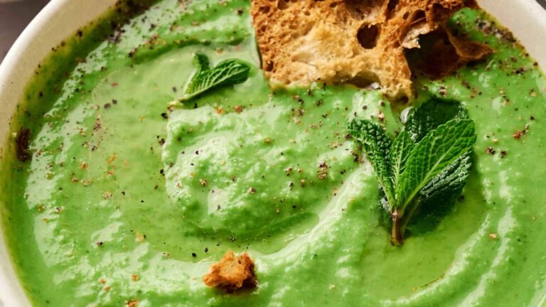 A bowl of thick green soup garnished with fresh mint leaves, cracked black pepper, and pieces of toasted bread.