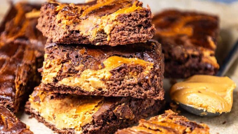 Three chocolate brownies with visible swirls of peanut butter are stacked on parchment paper, with a spoonful of peanut butter beside them.