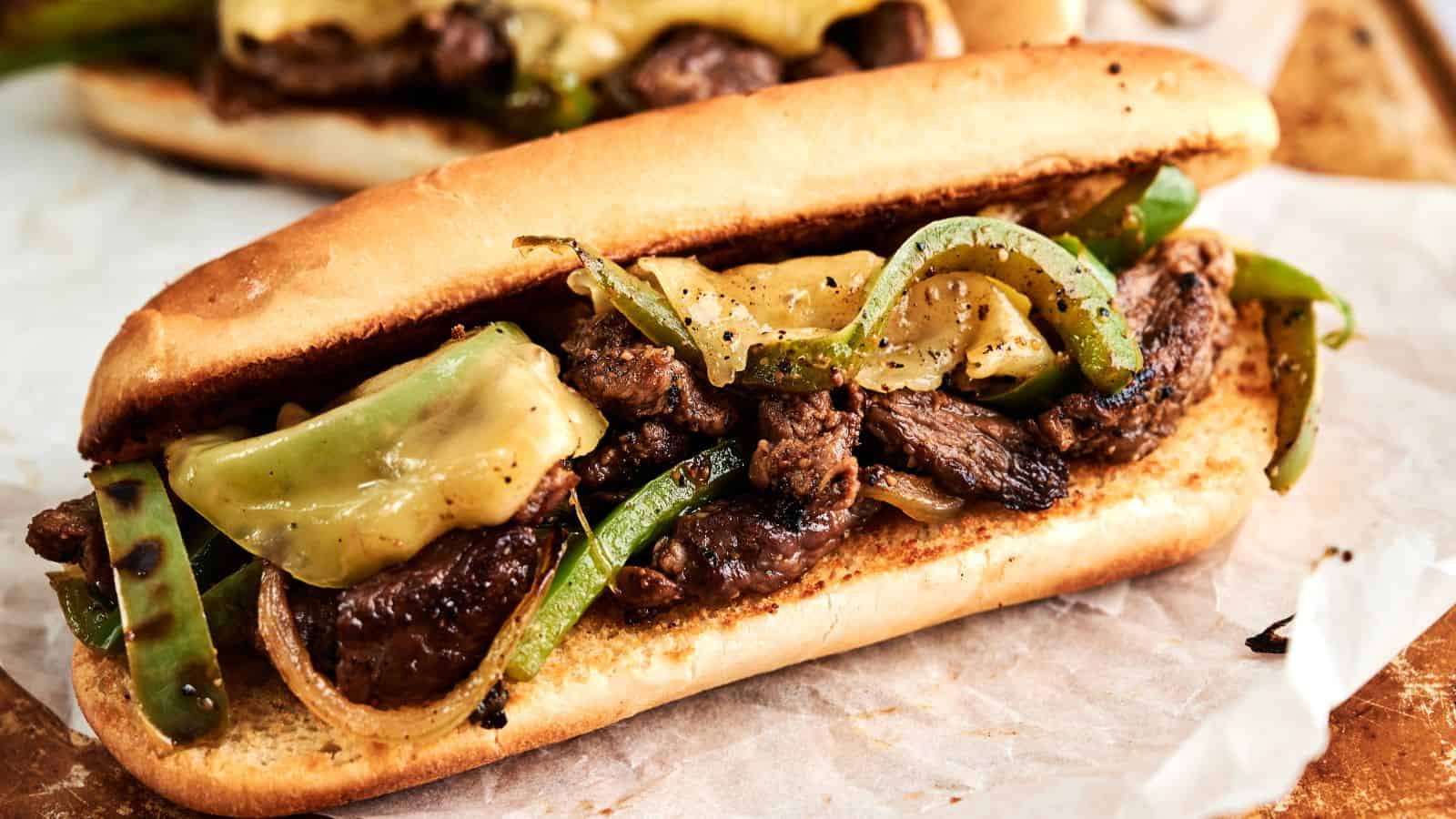A sandwich roll filled with grilled steak strips, melted cheese, green bell peppers, and onions on parchment paper.