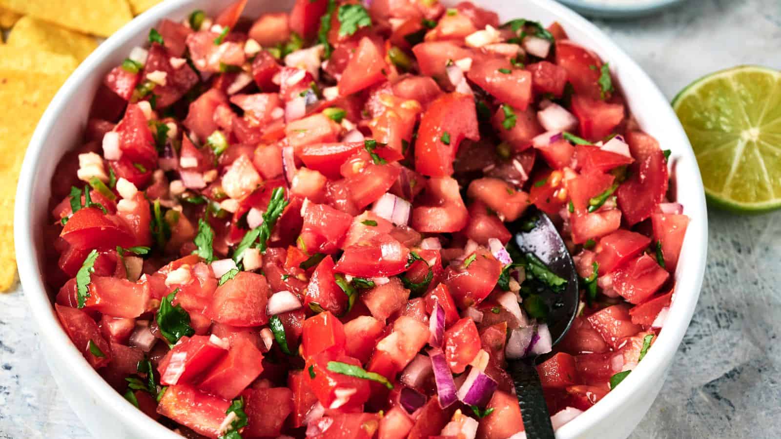 A bowl of pico de gallo with chopped tomatoes, onions, cilantro, and jalape&ntilde;os with a spoon inside, next to a lime and tortilla chips.