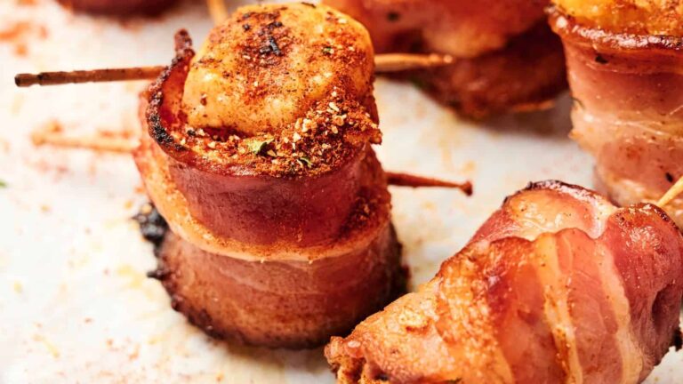 Close-up of scallops wrapped in bacon, secured with toothpicks, and seasoned, resting on a white baking sheet.