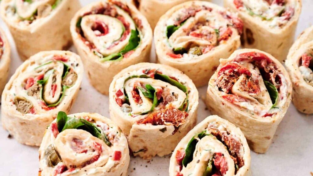 Several tortilla pinwheel sandwiches filled with spinach, roasted red peppers, and a creamy spread are arranged closely together on a white surface.