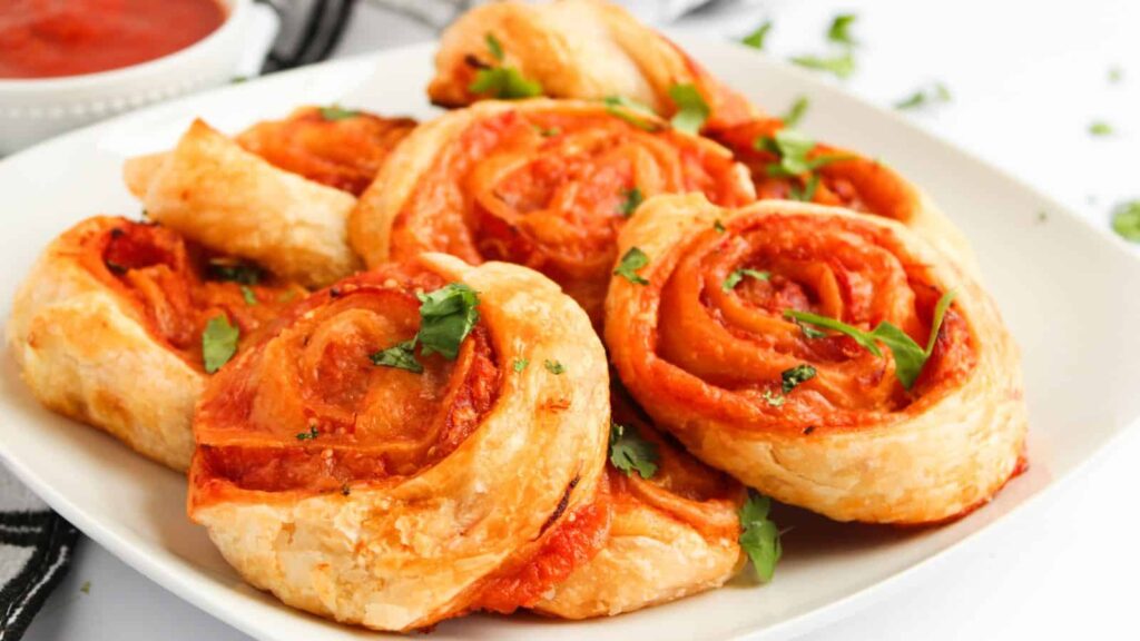 A white plate with several puff pastry pizza pinwheels topped with tomato sauce and garnished with chopped parsley.