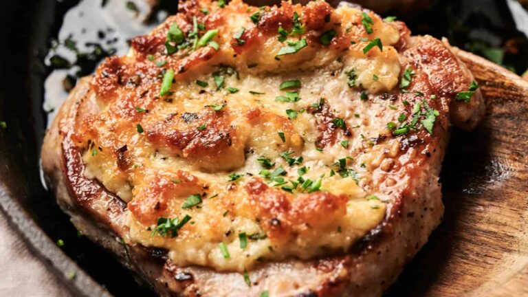 Close-up of a cooked pork chop topped with melted cheese and chopped herbs on a wooden serving spoon.