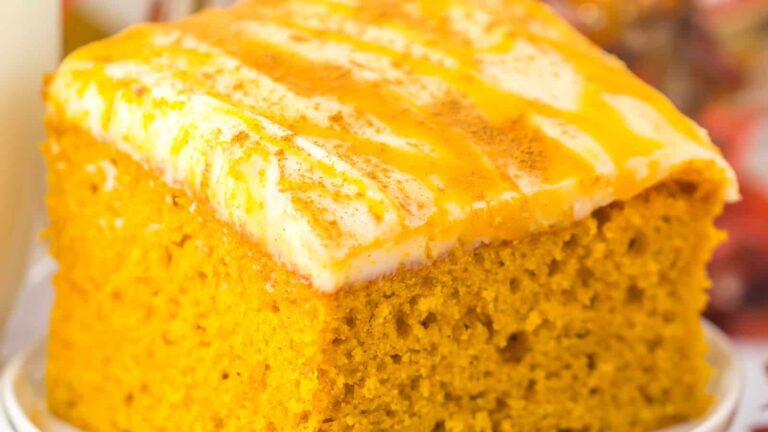 A close-up of a square slice of pumpkin cake topped with cream cheese frosting and drizzled with an orange glaze.