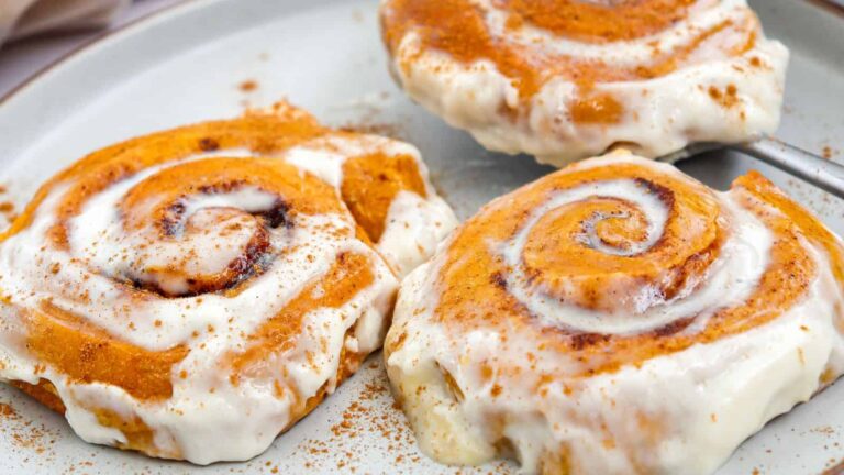 Three cinnamon rolls topped with creamy white icing and a sprinkle of cinnamon are arranged on a white plate.