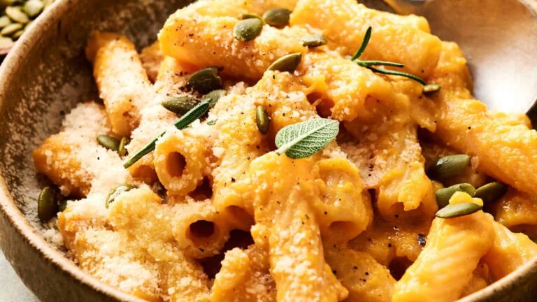 A bowl of rigatoni pasta coated in creamy sauce, topped with grated cheese, pumpkin seeds, and fresh herbs.