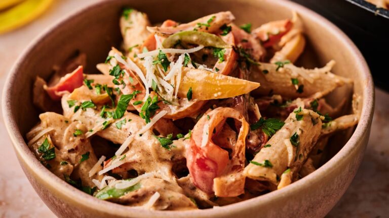 A bowl of penne pasta mixed with creamy sauce, sliced bell peppers, chopped parsley, and grated parmesan cheese.