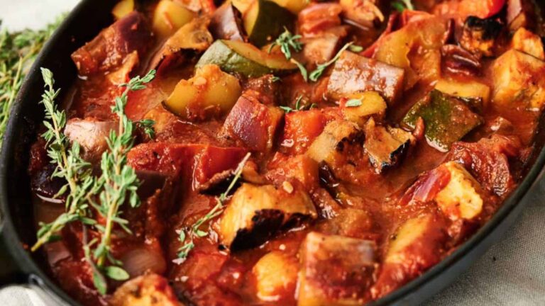 A close-up of a baked vegetable stew with chunks of zucchini, tomatoes, and eggplant, garnished with fresh thyme sprigs in a black dish.