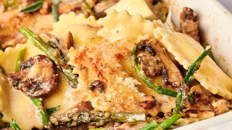 Close-up of baked ravioli with mushrooms, asparagus, and a golden-brown cheese topping in a white dish.