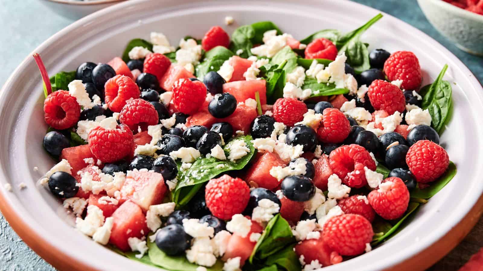 A salad with spinach leaves, watermelon cubes, blueberries, raspberries, and crumbled feta cheese in a white bowl.