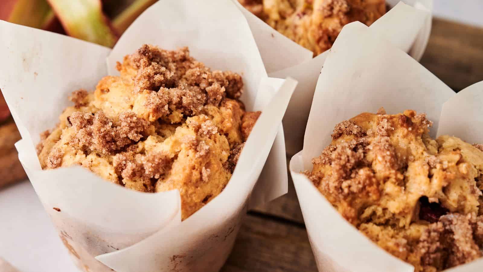 Close-up of three muffins with crumbly streusel topping, each wrapped in white parchment paper, arranged on a wooden surface.