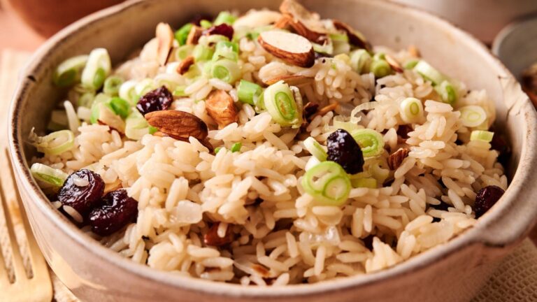 A bowl of cooked rice topped with sliced green onions, whole almonds, and dried cranberries.