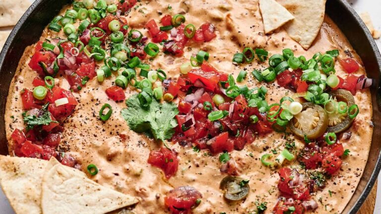 A skillet filled with cheesy queso dip, topped with chopped tomatoes, green onions, jalapeños, cilantro, and surrounded by tortilla chips.