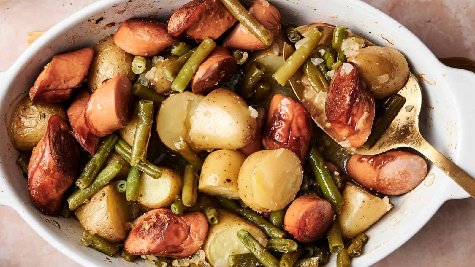 A baked dish containing sliced sausages, potatoes, and green beans in a white casserole dish with a serving spoon.