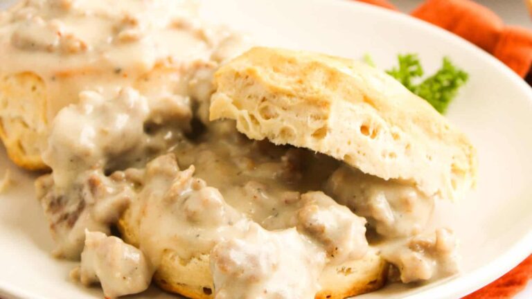 A plate with a biscuit topped with sausage gravy, with one biscuit half resting on top and a sprig of parsley on the side.