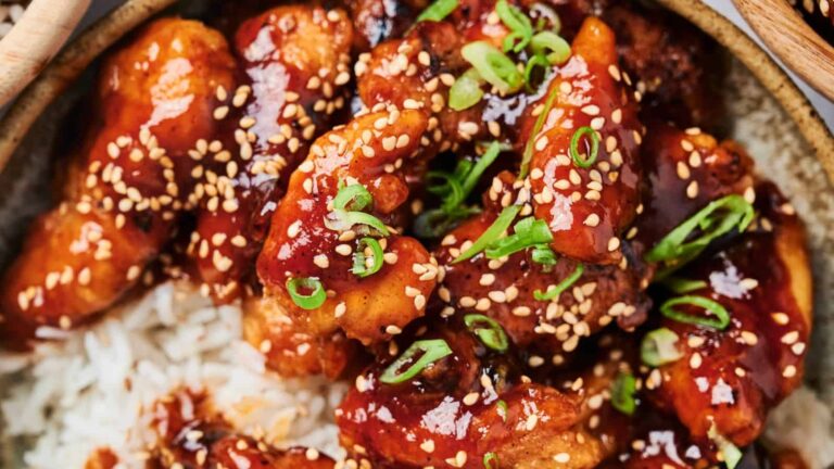 A close-up of glazed chicken pieces topped with sesame seeds and sliced green onions, served over white rice.