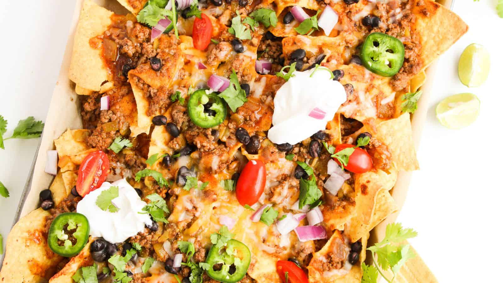 A platter of nachos topped with ground meat, melted cheese, sliced jalapeños, cherry tomatoes, black beans, red onions, cilantro, and dollops of sour cream. Lime wedges are on the side.