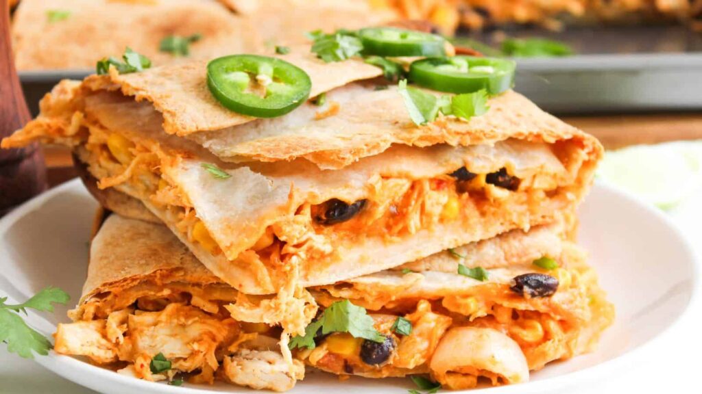Two pieces of stuffed quesadilla with chicken, black beans, cheese, and vegetables, garnished with sliced jalape&ntilde;os and cilantro, served on a white plate.