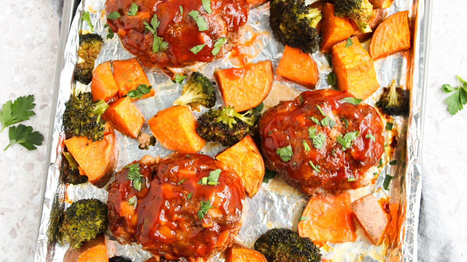 Three mini meatloafs topped with sauce are baked on a foil-lined tray with roasted sweet potato chunks and broccoli florets, garnished with chopped parsley.