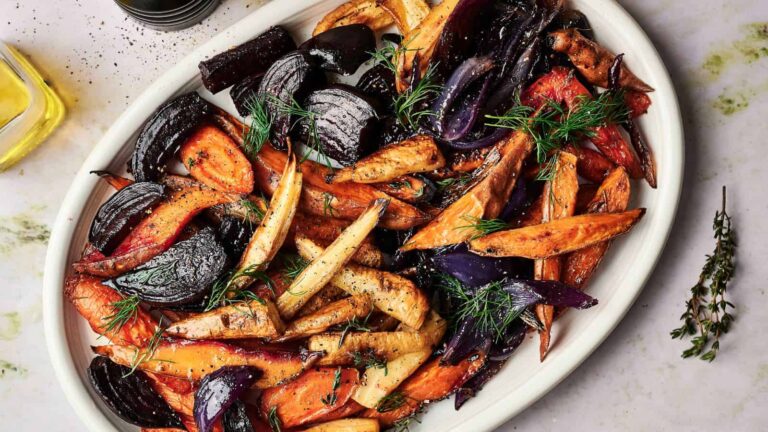 Oval platter of roasted root vegetables including carrots, parsnips, and red onions, garnished with fresh dill and black pepper on a light surface.