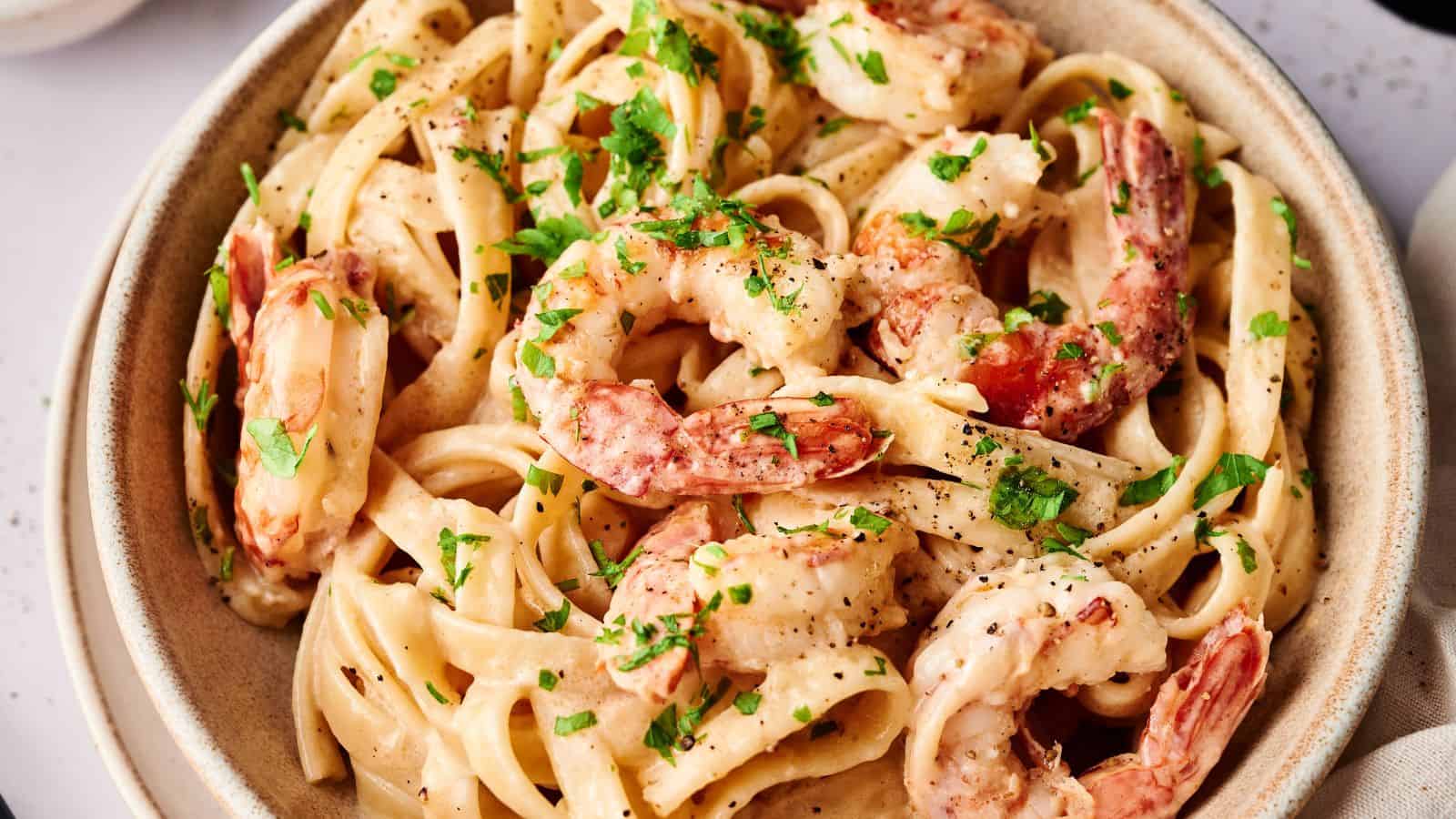 A bowl of fettuccine pasta with shrimp in a creamy sauce, garnished with chopped parsley and black pepper.