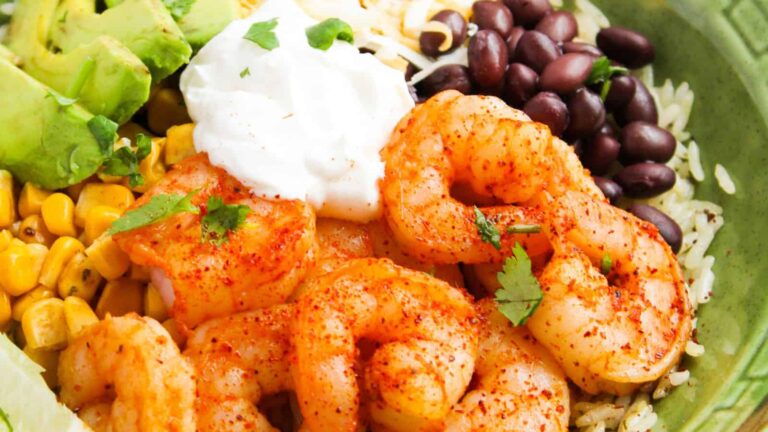 A bowl with seasoned shrimp, black beans, corn, avocado chunks, shredded cheese, white rice, and a dollop of sour cream, garnished with cilantro.