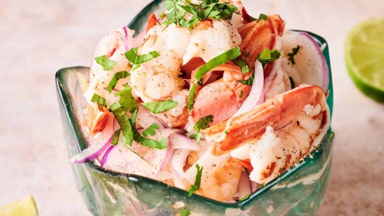A glass bowl filled with shrimp, sliced red onions, and chopped herbs, with a lime half in the background.