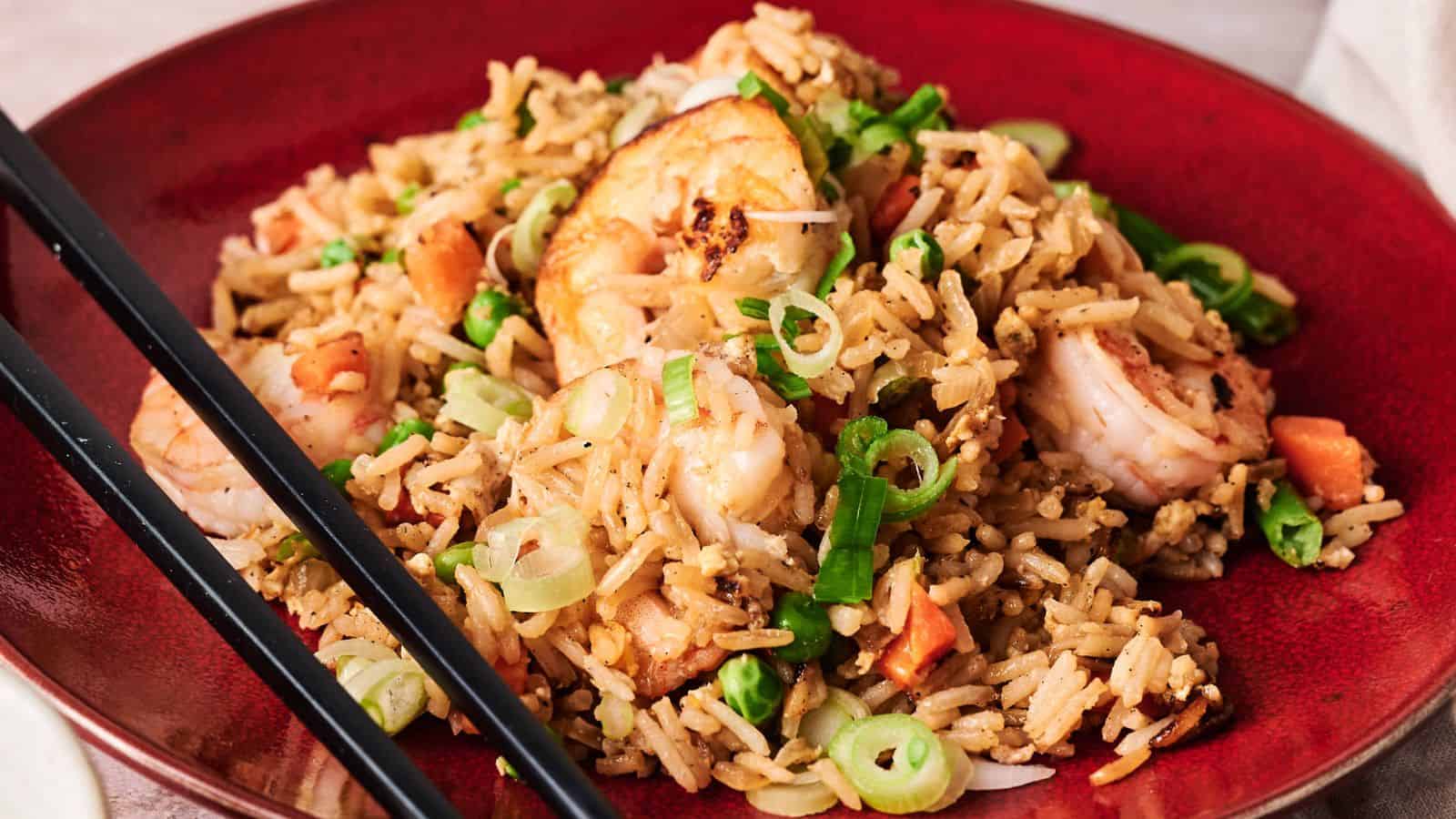 A red plate of shrimp fried rice garnished with chopped green onions, with black chopsticks resting on the side.