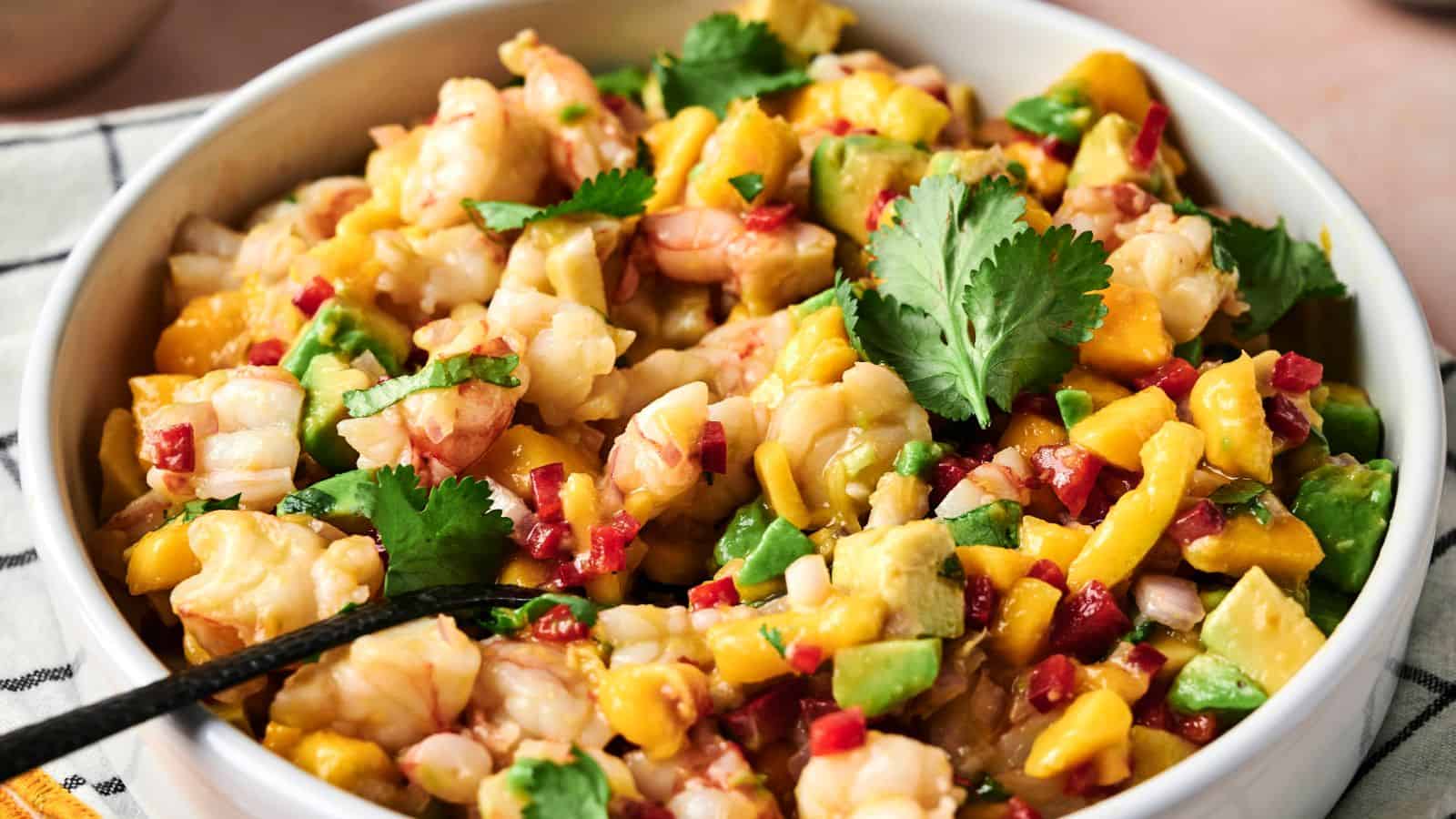 A bowl of shrimp ceviche with diced mango, avocado, red onion, red pepper, and cilantro, with a spoon inside.