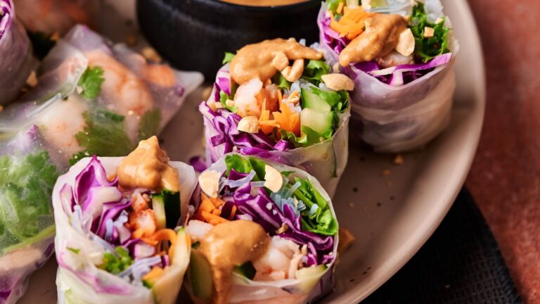 Fresh spring rolls filled with shrimp, lettuce, purple cabbage, carrots, and herbs, topped with peanut sauce and chopped peanuts, served on a plate.
