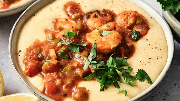 A bowl of creamy grits topped with shrimp, tomato-based sauce, diced vegetables, and garnished with fresh parsley.