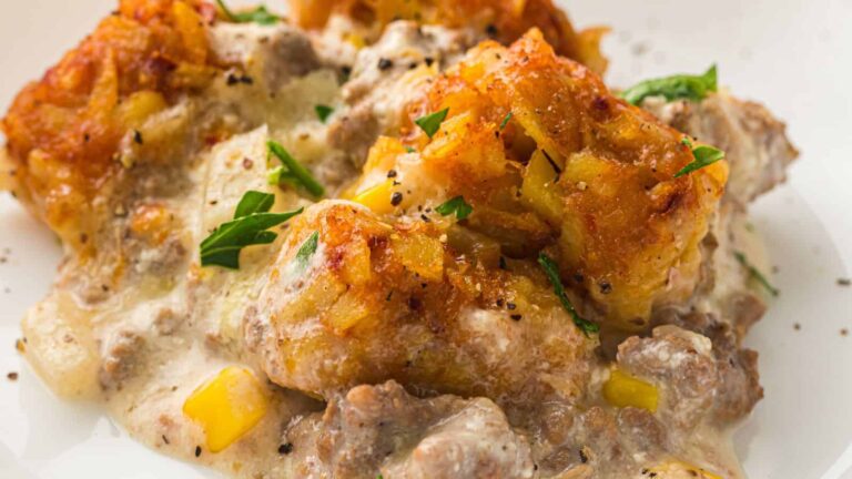 A close-up of a cheesy tater tot casserole with ground beef, yellow bell peppers, creamy sauce, and parsley garnish on a white plate.