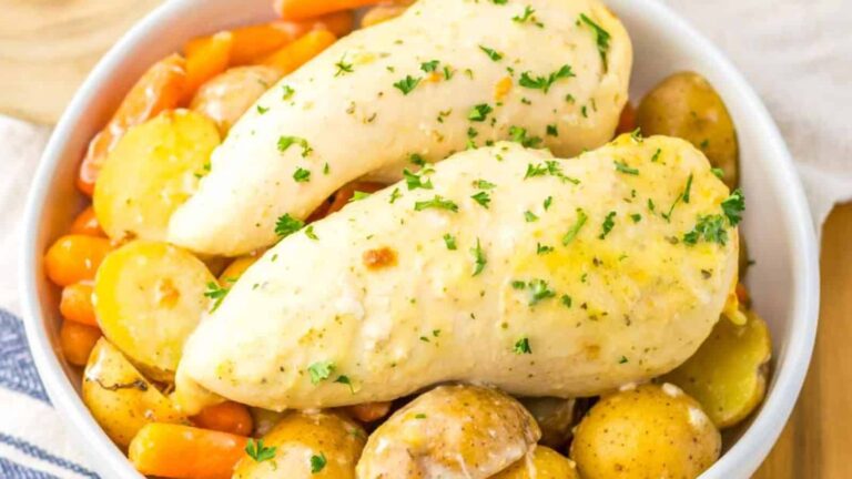 Two cooked chicken breasts topped with chopped herbs served over baby potatoes and carrots in a white bowl.