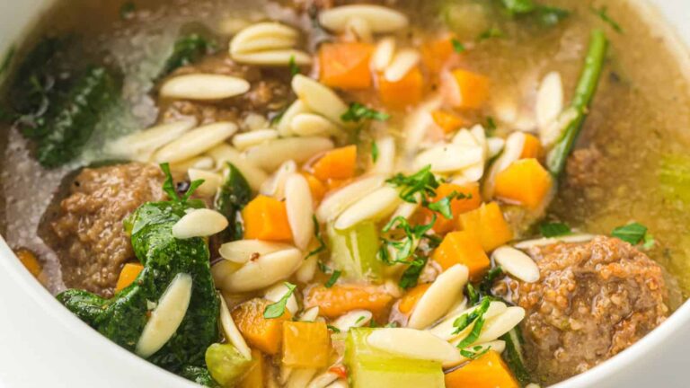 A close-up of a bowl of soup with meatballs, diced carrots, celery, spinach, and orzo pasta in broth, garnished with chopped herbs.
