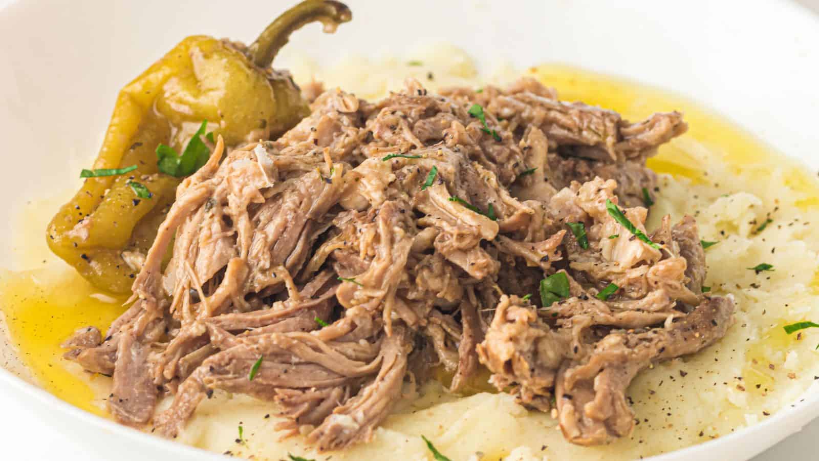 Shredded roast beef served over mashed potatoes with a pepper and a light drizzle of gravy, garnished with chopped herbs.