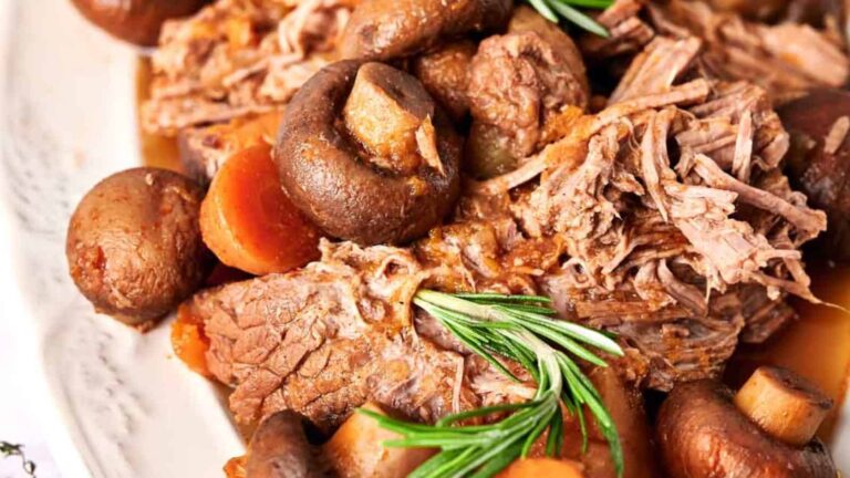 Close-up of a pot roast with shredded beef, whole mushrooms, carrot slices, and a sprig of fresh rosemary on a white plate.