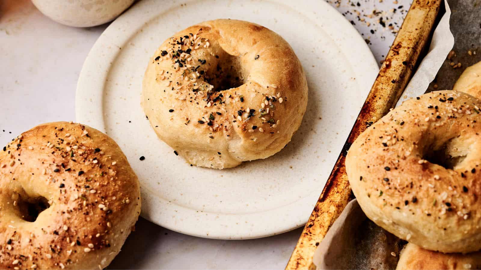 A plate with a bagel seasoned with seeds sits next to a baking tray holding more bagels, also topped with seeds.