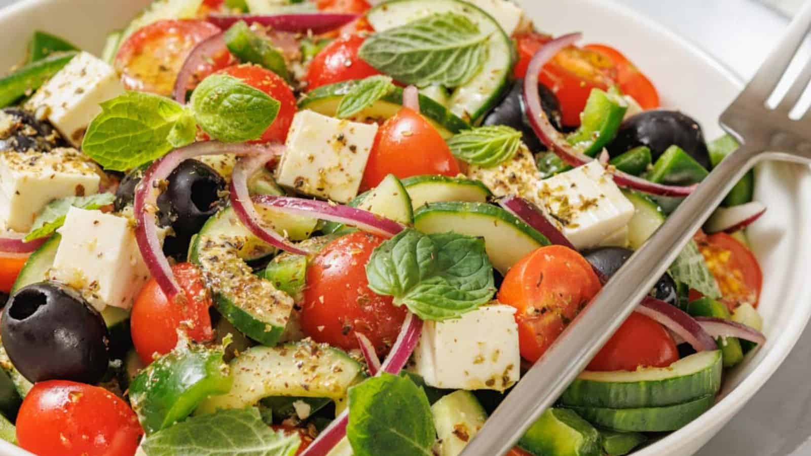 A bowl of Greek salad with cherry tomatoes, cucumber slices, red onion, black olives, feta cheese, and fresh mint leaves, sprinkled with herbs. A fork rests on the side.