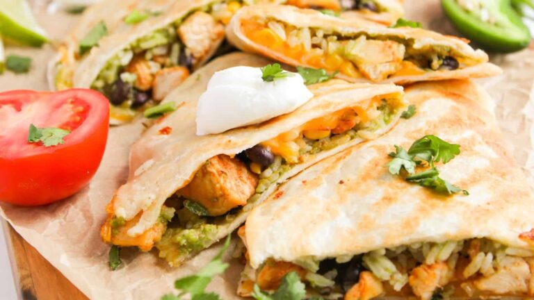 Three quesadilla wedges filled with chicken, rice, beans, and cheese, topped with a dollop of sour cream and garnished with cilantro, next to sliced tomato and jalapeño.