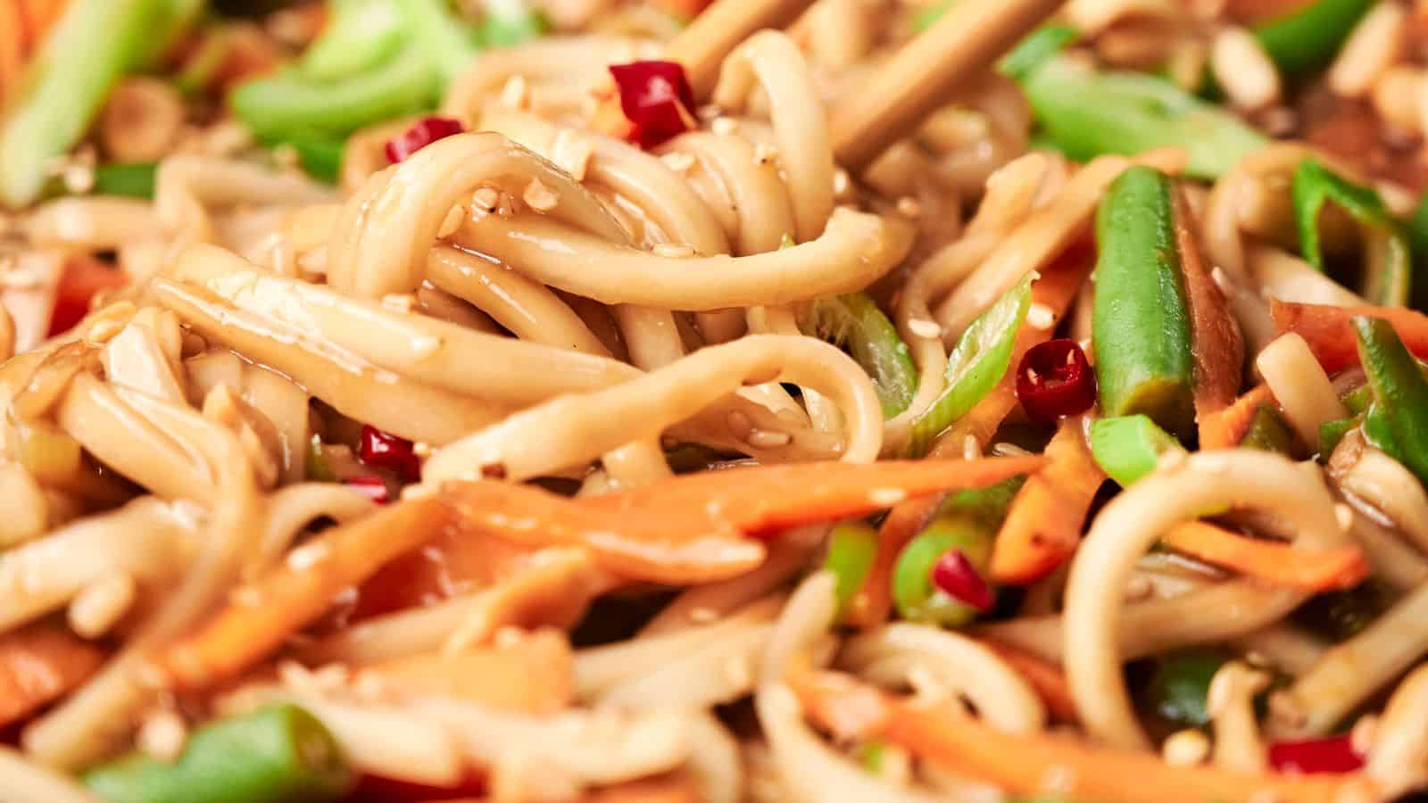 Close-up of stir-fried noodles with vegetables, including carrots, green beans, and chopped red peppers, garnished with sesame seeds. Chopsticks are picking up some noodles.