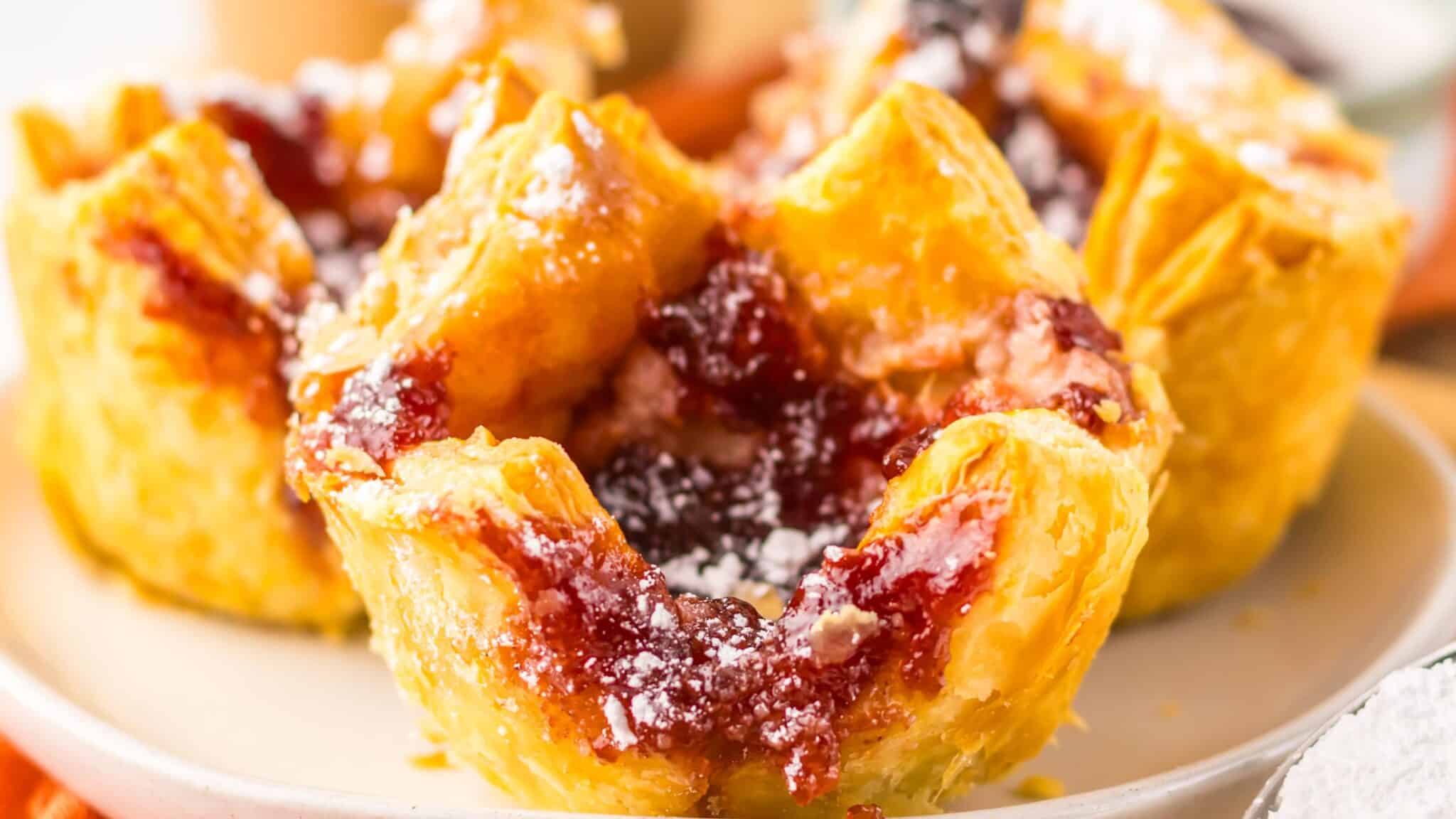 Close-up of flaky pastries filled with red fruit jam, sprinkled with powdered sugar, on a white plate.